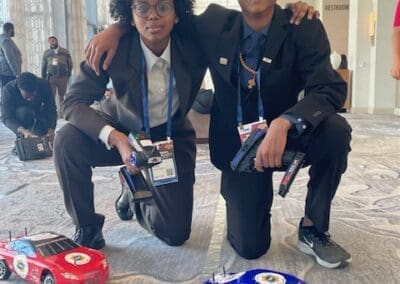 MKYF Two individuals in business attire crouching beside model racing cars at an indoor event.