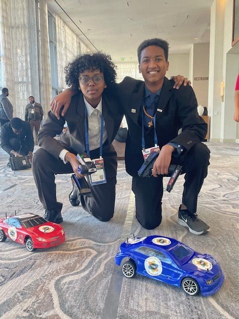 MKYF Two individuals in business attire crouching beside model racing cars at an indoor event.