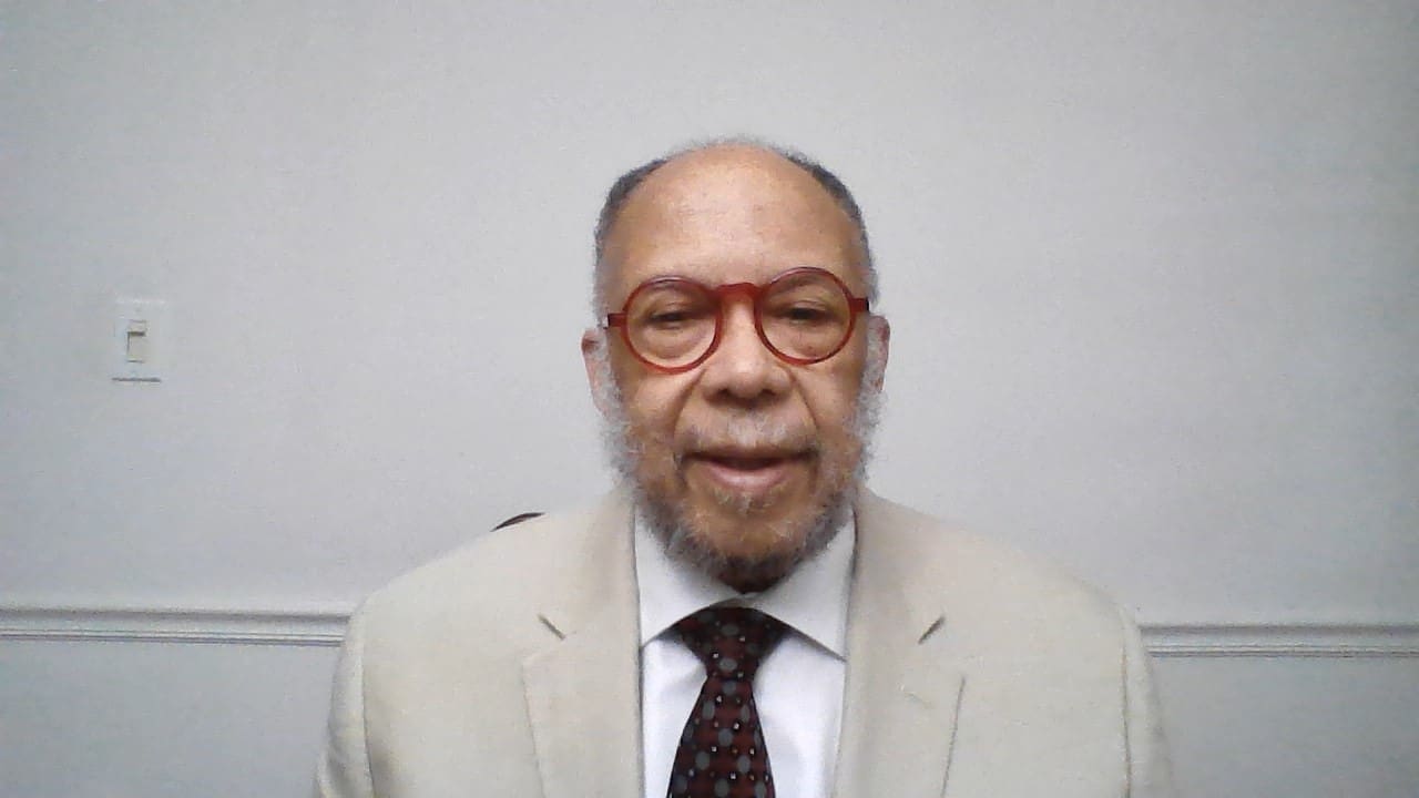 MKYF A man with gray hair and beard, wearing red round glasses, a beige suit jacket, white shirt, and dark patterned tie, stands in front of a light gray background.