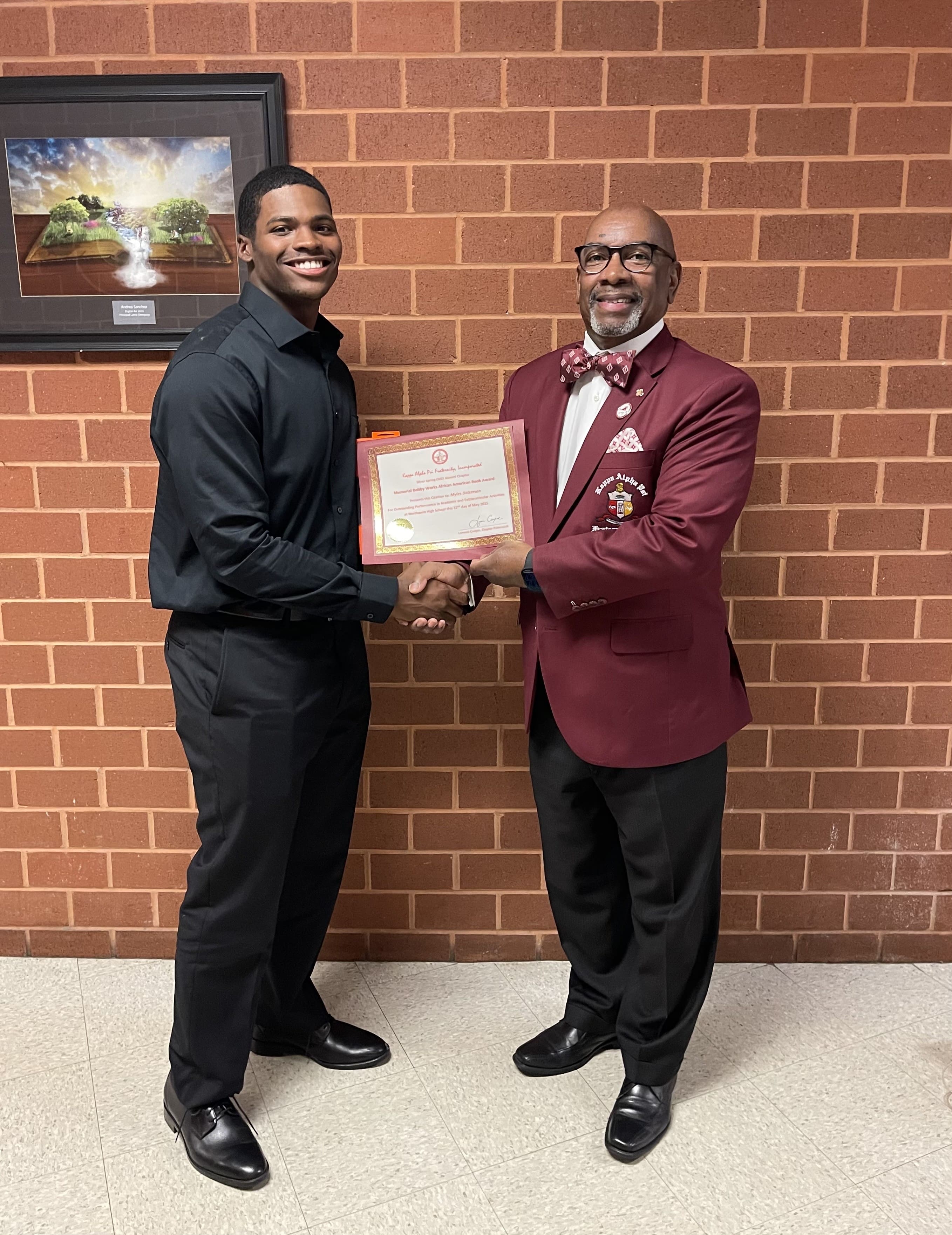 MKYF Two men shake hands while one presents a framed certificate to the other. They stand in front of a brick wall with a framed picture above them.