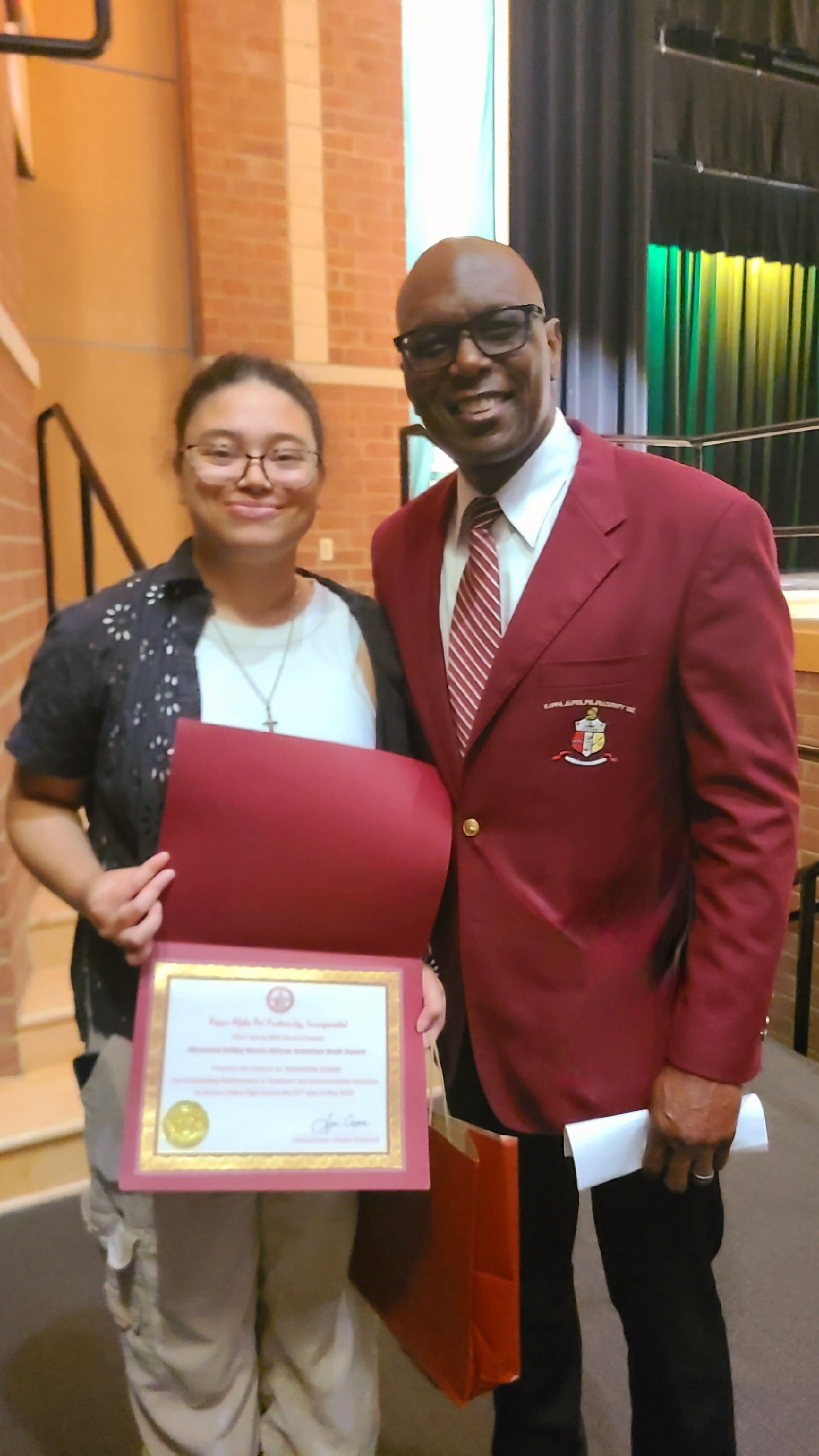 MKYF A person holds a certificate while standing next to a smiling man in a maroon blazer and striped tie on a stage with brick walls and curtains in the background.