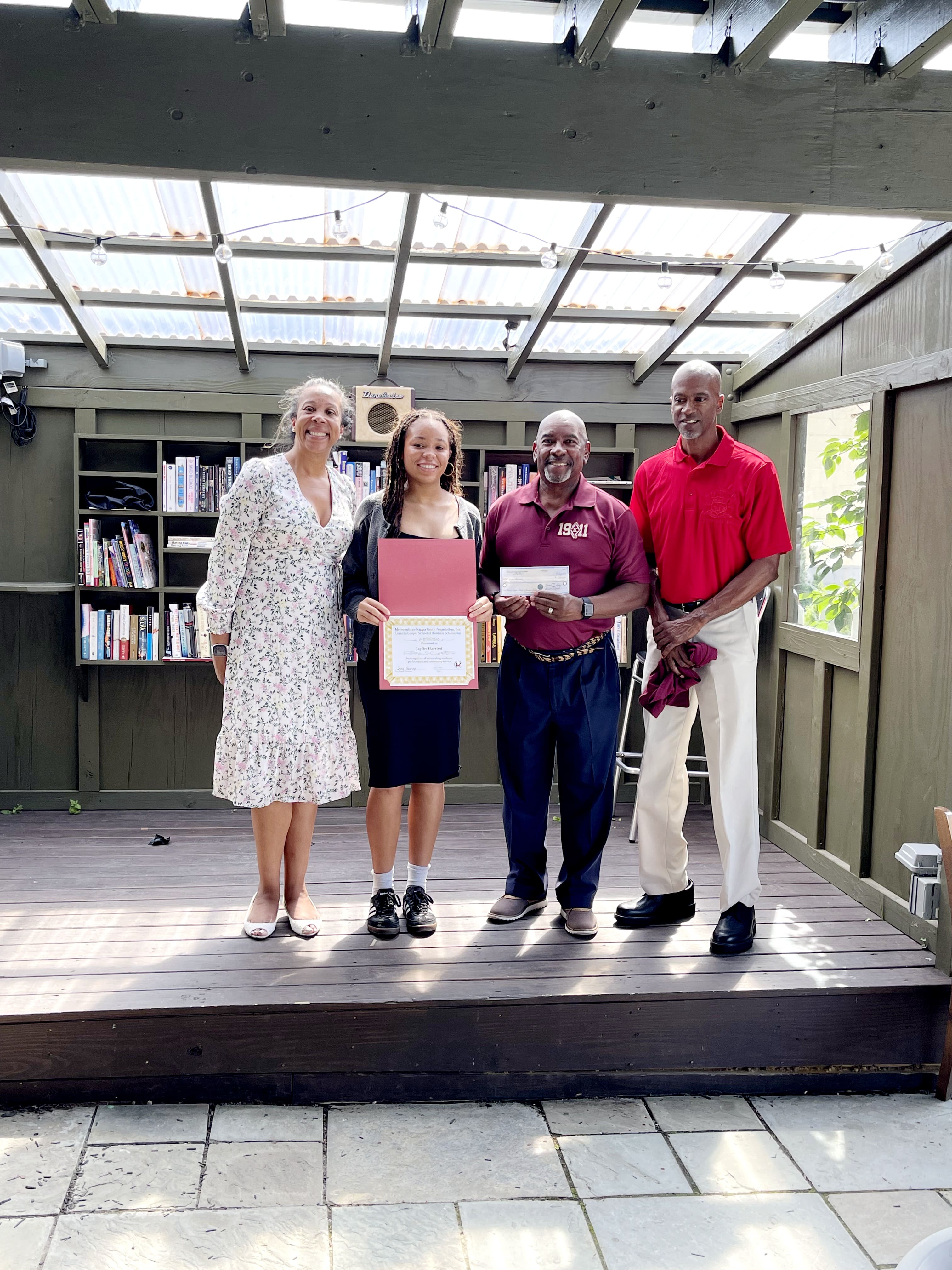 MKYF Four adults stand on a wooden stage; the person second from left holds a certificate and a folder. Shelves of books are in the background.
