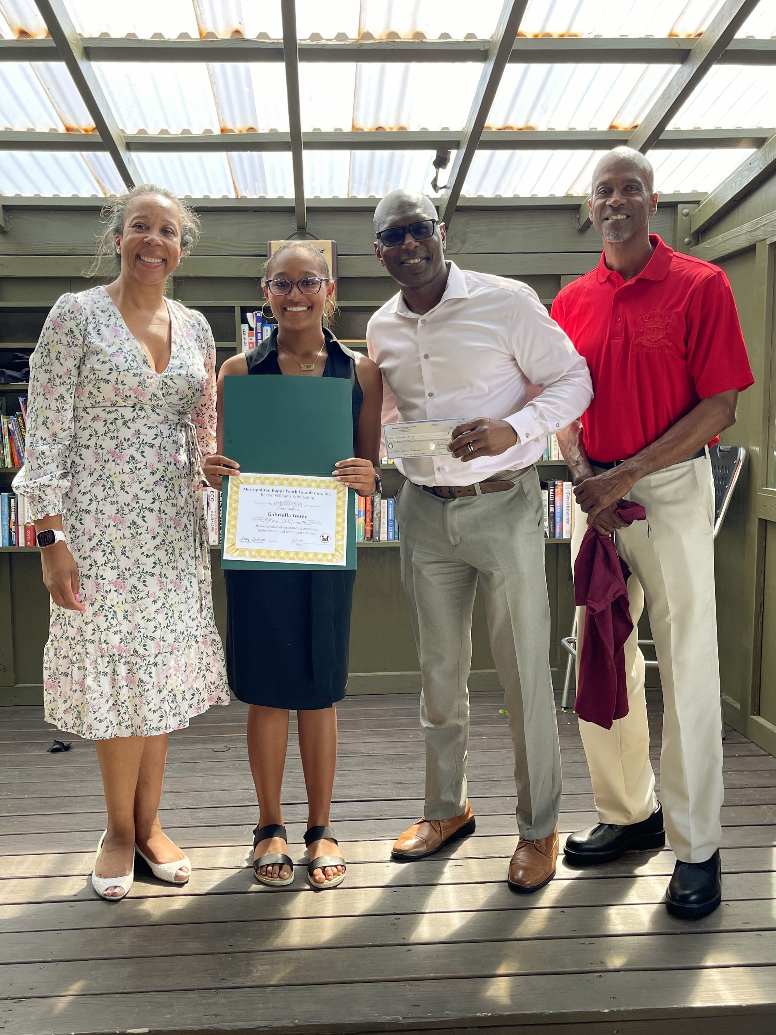 MKYF Four people stand together on a wooden deck; one young person holds a certificate and another adult holds a check. Shelves with books are visible in the background.