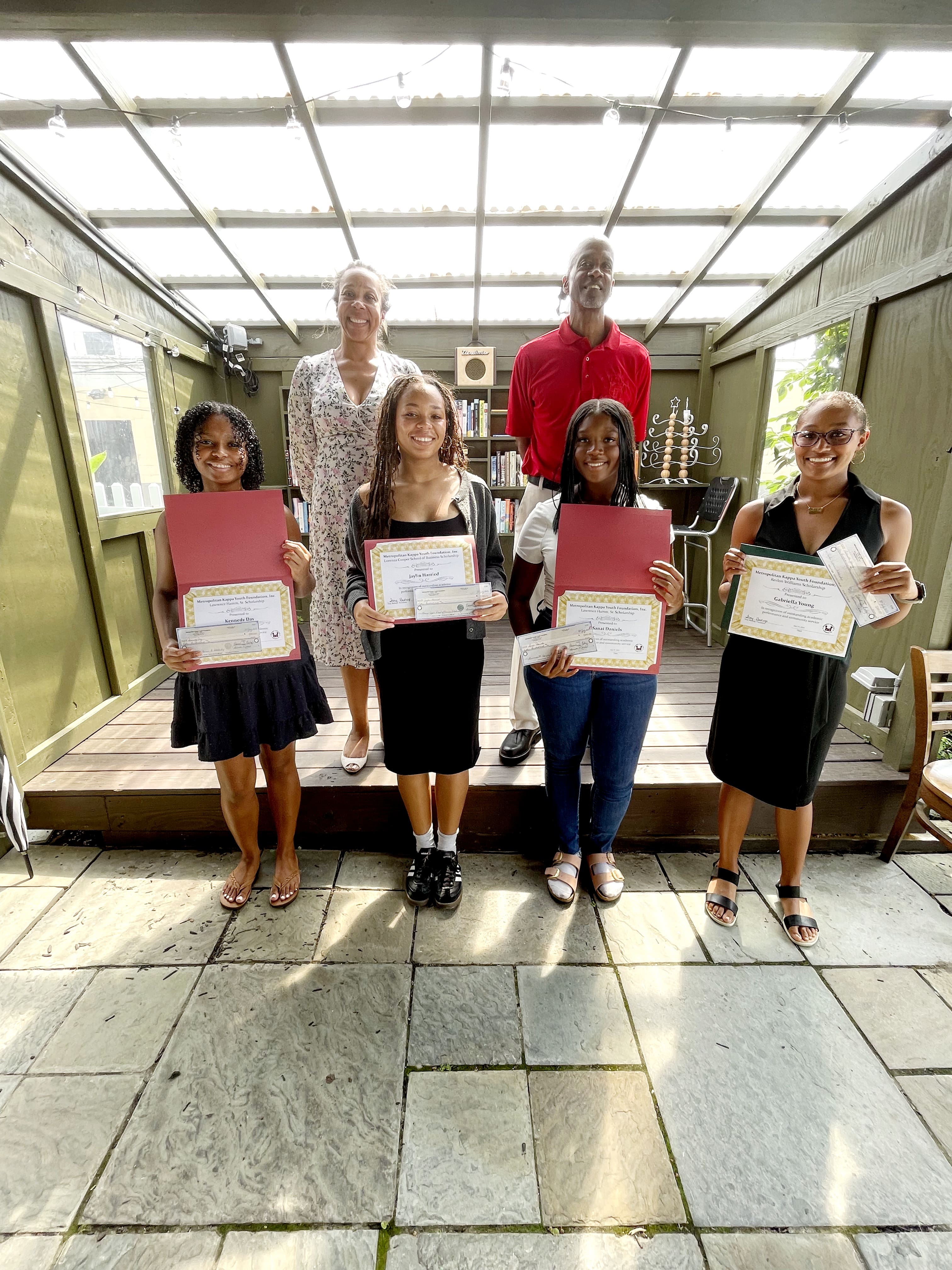 MKYF Four young women hold certificates and folders while standing in front of two adults on a small stage under a glass roof in an outdoor setting.