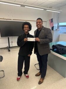 MKYF Two men standing in a classroom; one is handing a check to the other. Both are smiling, and there is a large monitor and an American flag in the background.