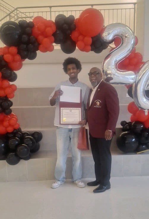 MKYF Two men pose together; one holds a certificate. They stand in front of a arch made of red and black balloons and large silver "24" balloons.