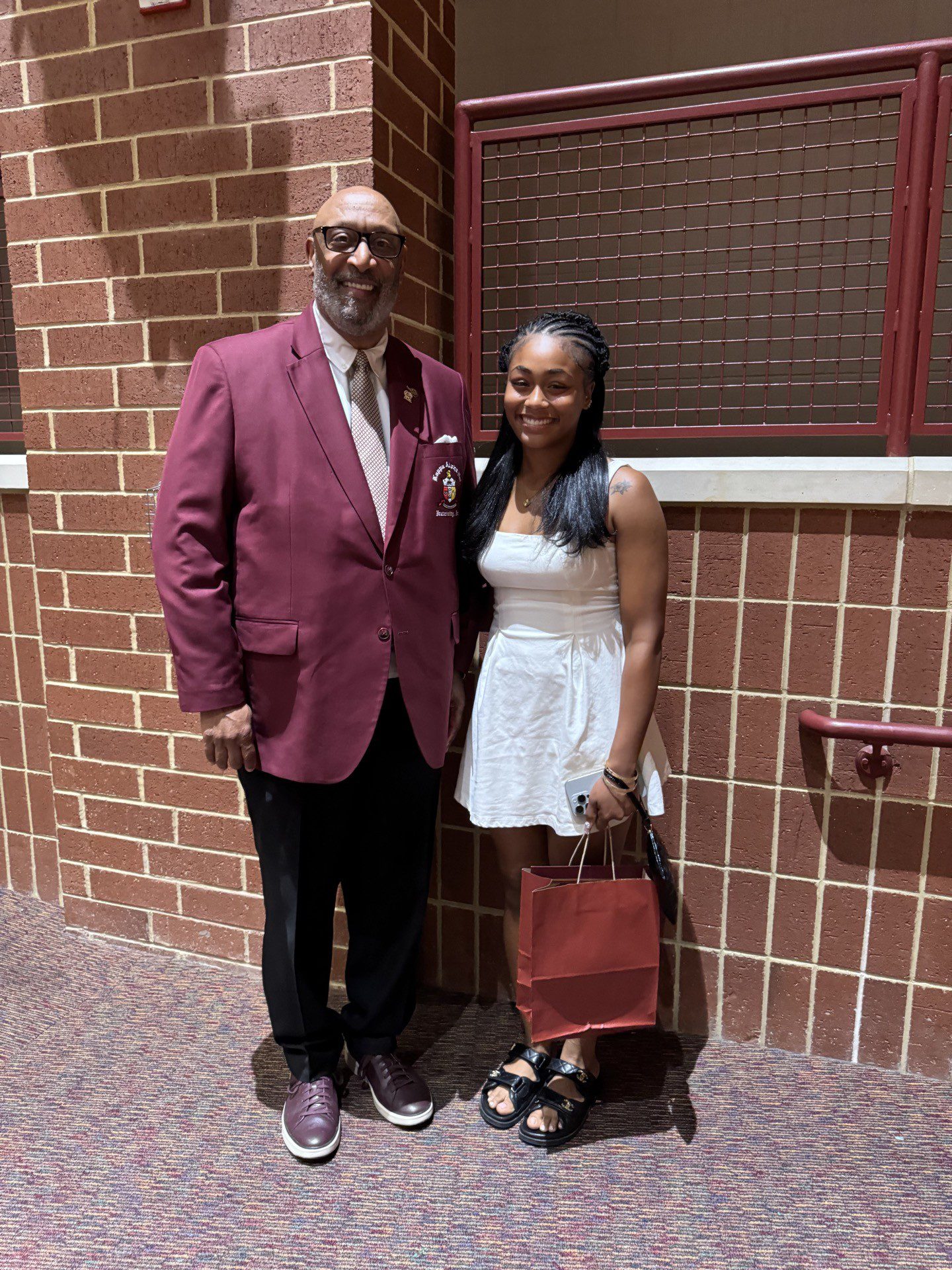 MKYF A man in a maroon blazer stands next to a woman in a white dress, both smiling, in front of a brick wall indoors. The woman is holding a red gift bag.